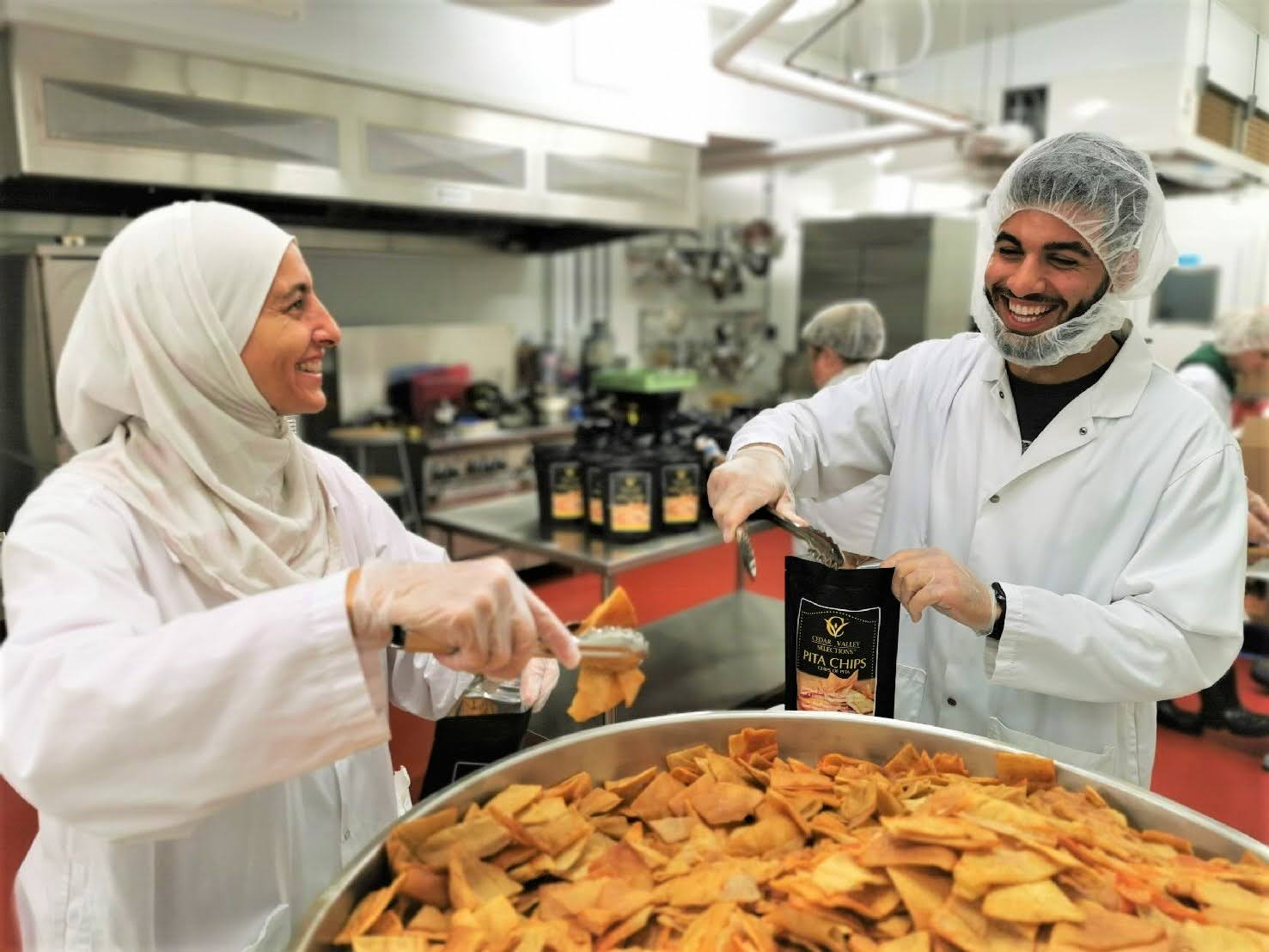 Two workers smiling near large pot of food in kitchen facility.